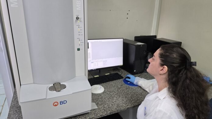 Laboratory technician in a white coat and gloves sits at a desk, monitoring a BD instrument and a computer screen.