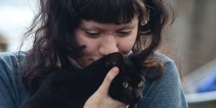 Person with dark hair gently cradling a black cat, both looking downward; cat's yellow eyes visible.