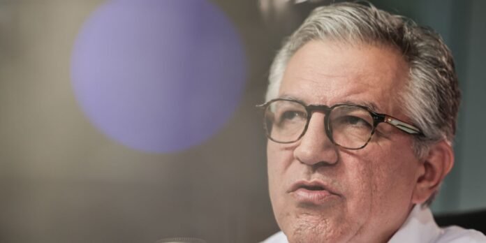 Close-up of an older man with gray hair and tortoiseshell glasses, looking to the right, with a purple bokeh circle in the background.