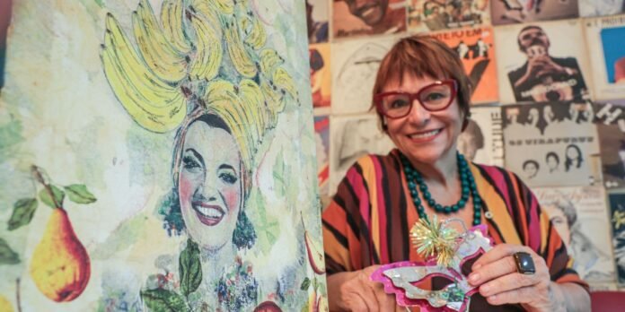 Smiling woman with red glasses and striped colorful blouse holding a decorative mask against a wall of vintage posters.