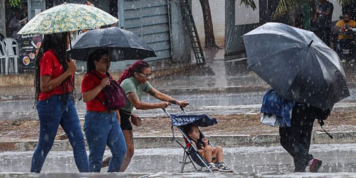 Group of people walking in heavy rain under umbrellas; a child sits in a stroller being pushed along the wet sidewalk.
