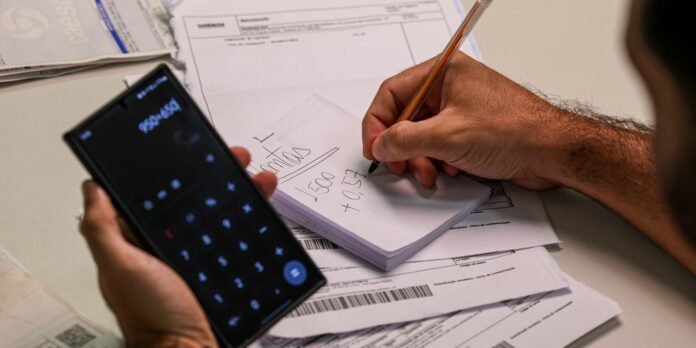 Person holding a smartphone with a calculator app while writing numbers in a notebook over stacked bills and invoices on a desk.