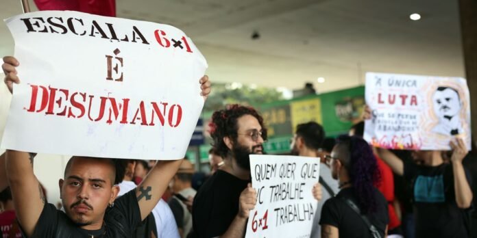 Protester raises a large white sign with red letters reading 'ESCALA 6x1 E DESUMANO' at a crowded demonstration in a public space.