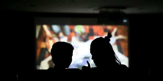 Two people in silhouette watch a colorful projection on a large screen in a dark room.