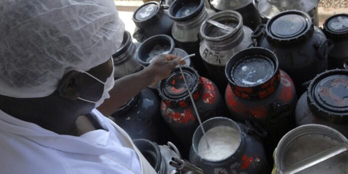 Person in protective suit, mask, and hairnet stirs a white substance from a large open container among red-and-black paint drums.