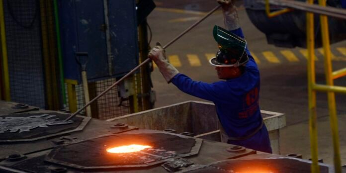 Factory worker in blue coveralls and a face shield uses a long rod to move near a glowing molten metal pool in an industrial foundry.