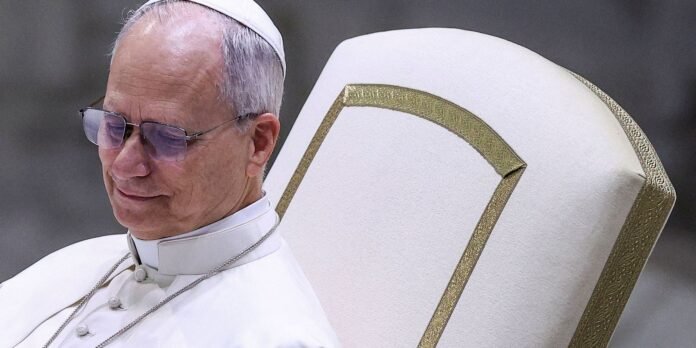 Senior man in white clerical robes and a skullcap sits on a large white chair with gold trim and a regal demeanor.