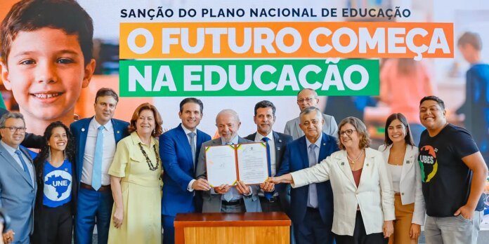 Group of officials and supporters pose with an open document during a signing ceremony, a large banner reads 'O FUTURO COMEÇA NA EDUCAÇÃO' in the background.