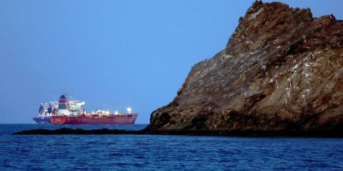 Large red cargo ship sailing near a steep rocky cliff by calm blue water at dusk/dark blue sky nearby shore