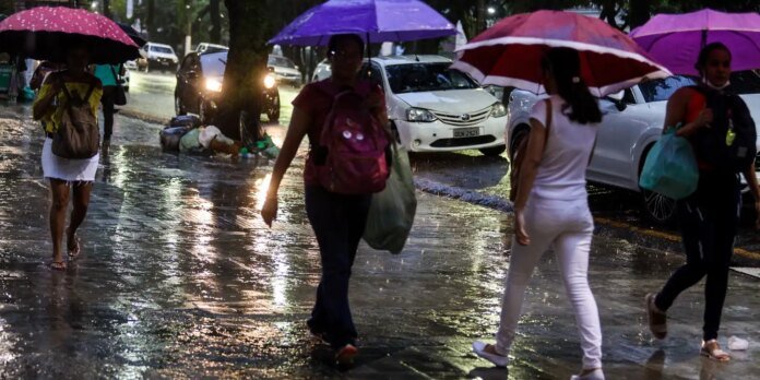 People with colorful umbrellas walking on a wet city street at dusk, car headlights in the background and reflections on the pavement.