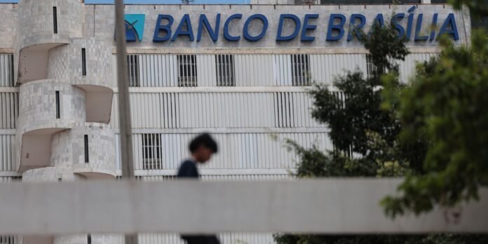 Facade of Banco de Brasília building with blue sign on a stone wall and vertical white louvers; a blurred person walks in foreground.