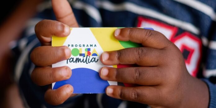 Hands hold a colorful Bolsa Família program card against a blurred striped shirt background