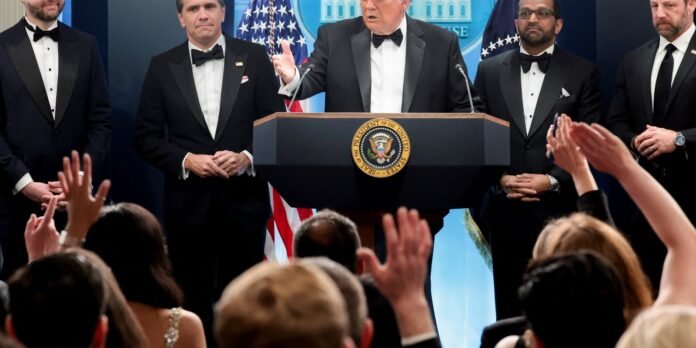 President at a podium with the Seal of the United States, flanked by men in tuxedos, addressing a crowd as audience members raise their hands in applause.
