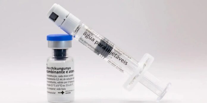 A syringe attached to a small vial with a blue cap, ready for injection, on a pale gradient background (medical scene). Iden­tifiable vaccine context, though label text is blurred.