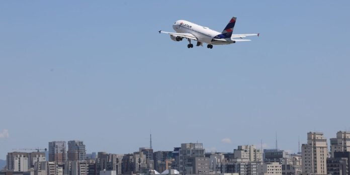 A white LATAM airplane taking off over a city skyline against a clear blue sky, with its landing gear down.