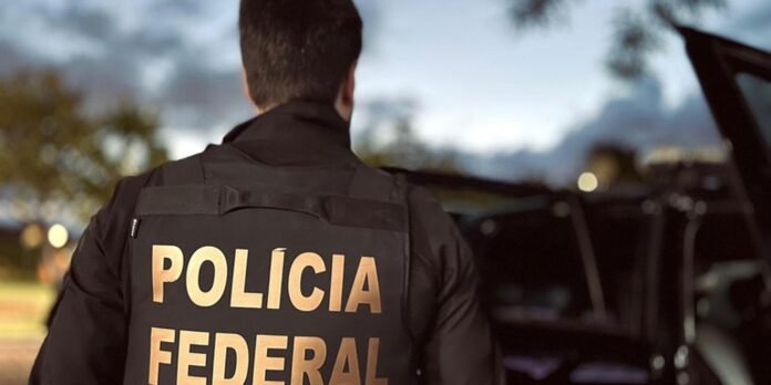 Back view of a police officer wearing a black vest labeled 'POLICIA FEDERAL' standing near an open car at dusk with blurred lights in the background (informative)