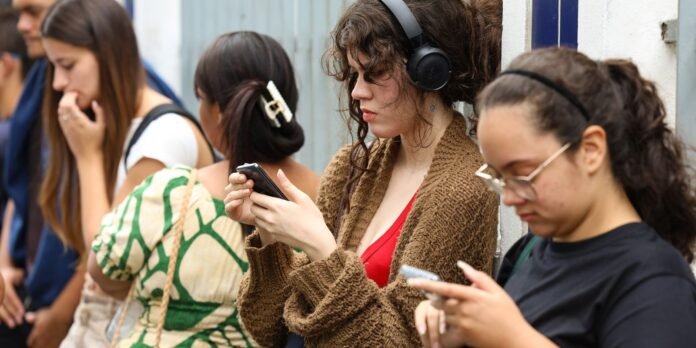 A woman in a brown knit cardigan with headphones looks down at her phone while others in line wait behind her.
