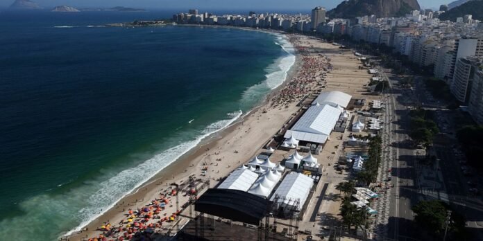 Aerial view of a busy beach with white festival tents, a large stage, and crowds along the shoreline beside tall city buildings.