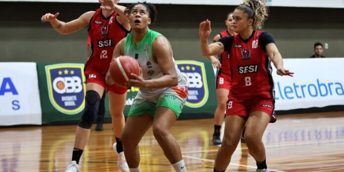 Woman in green drives with ball while two players in red defend on a basketball court close by banners.