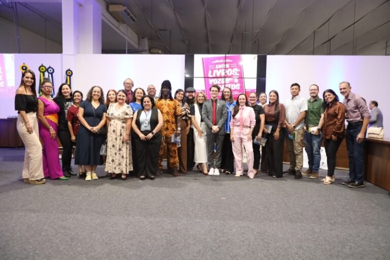 Group of about 25 diverse adults standing in a line for a group photo at a conference or event, indoors with white panels, purple lighting, and a pink banner in the background; attendees wear a mix of business casual and cultural attire.