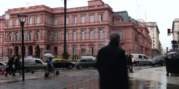 Urban street scene with a large pink historic building, wet pavement, and pedestrians including someone with an umbrella.
