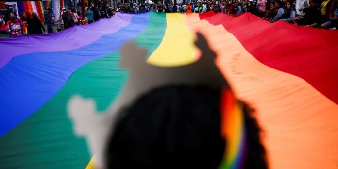 Wide rainbow Pride flag stretched across a crowded street, with people and colorful banners in the background and a blurred silhouette cast on the fabric.