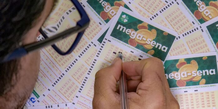 Close-up of a person wearing glasses, holding a pen and marking numbers on Mega-Sena lottery tickets spread on a table.