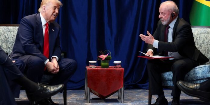 Two men in dark suits sit facing each other and converse, a small red table with a flower arrangement between them behind a blue curtain.