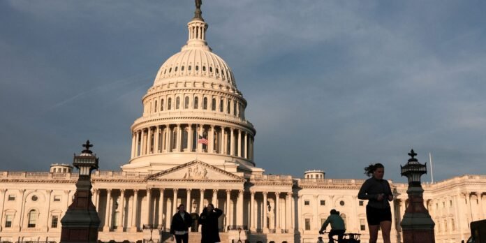 Wide view of the U.S. Capitol dome with pedestrians and a cyclist in the foreground near ornate street lamps.