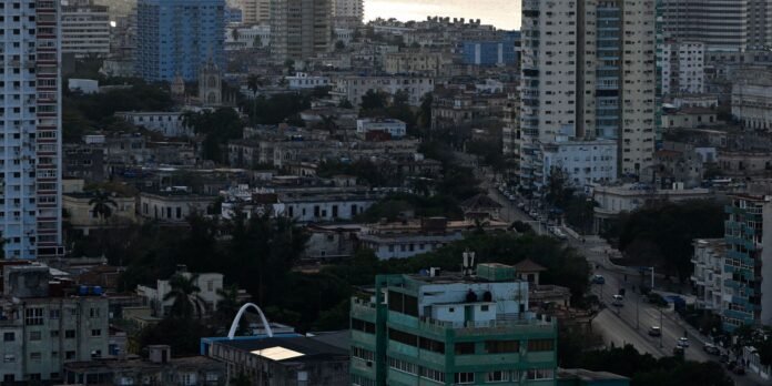 Dense urban skyline with low-rise buildings and tall towers, a street with cars, and a distant sunset over the water.