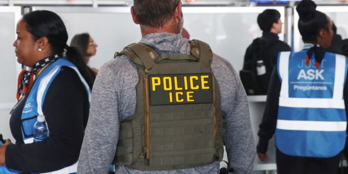 Back view of a man in a green tactical vest with a bold POLICE ICE patch, standing at a counter among others in blue vests.