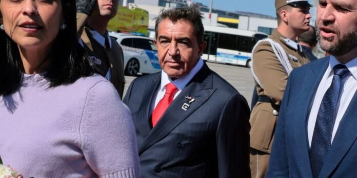Man in a dark suit with a red tie and lapel pins stands among suited colleagues and uniformed guards at an outdoor event, looking ahead.
