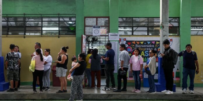 People line up outside a green-yellow school building, waiting to enter for class with a colorful banner in the background.