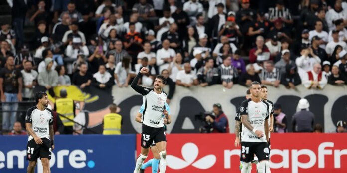 Soccer players in white and black uniforms celebrate on the pitch while a cheering crowd looks on from the stands.