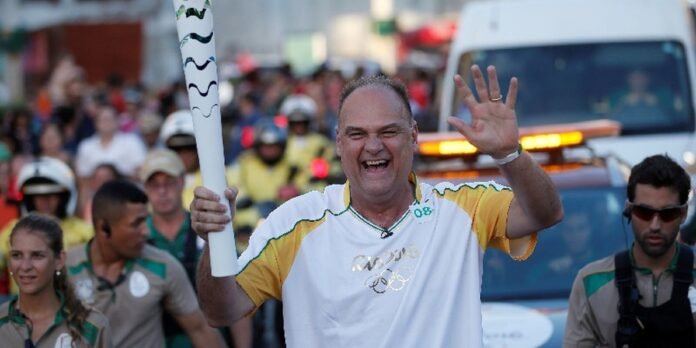 Smiling man waves while holding a white Olympic torch during a street parade, with a cheering crowd behind him.