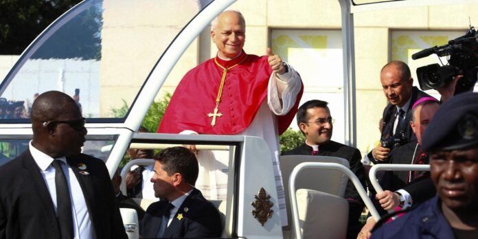 Religious leader in red robes giving a thumbs up from a vehicle, surrounded by security and clergy as cameras film the moment.