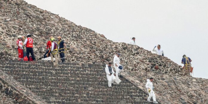 Rescue workers and investigators in white protective suits and red vests on a stone pyramid-like structure, coordinating on a rocky hillside during a relief operation.