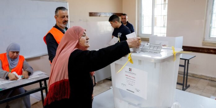 Older woman in a pink hijab casts a ballot into a white ballot box as volunteers in orange vests supervise a polling station environment.
