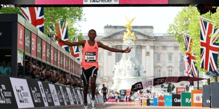 Male runner wearing a pink athletic top and black shorts crosses the finish line with arms outstretched as crowds cheer along a barrier-lined street in London, Union Jack flags visible.