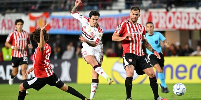 Soccer players in red-and-white striped kits and white kit contest the ball mid‑kick on a glossy green pitch, with a referee in blue and a yellow advertising banner in the background.