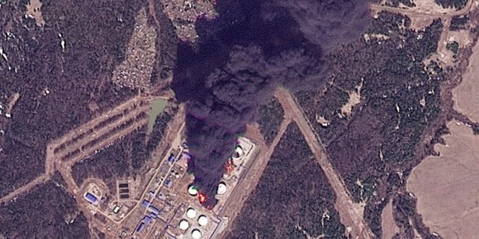 Aerial view of an industrial complex with circular storage tanks and a large black smoke plume rising from the center, near rugged terrain.