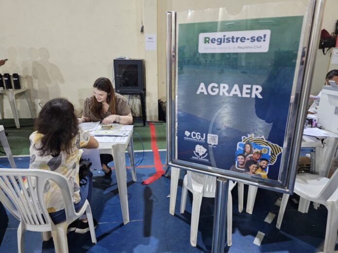 Two women at a desk filling forms at a civil registry booth in a gym, with a 'Registre-se!' poster nearby.