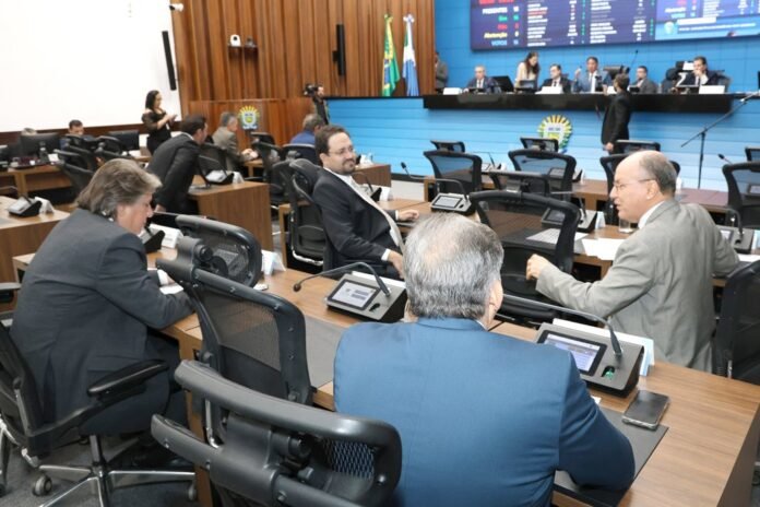Council chamber with officials seated at desks and a panel at the front on a blue backdrop with flags in view.