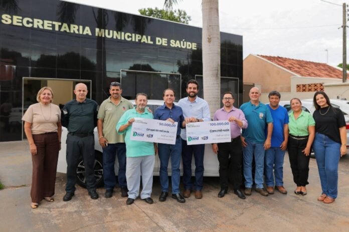 Group of people standing in front of a municipal health department building, holding two large ceremonial checks for 50,000 and 100,000 in a donation presentation gesture.