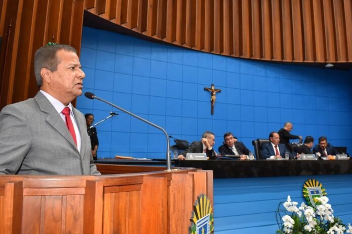 Man in a gray suit and red tie speaks at a wooden podium in a formal meeting hall with a blue wall and crucifix behind.