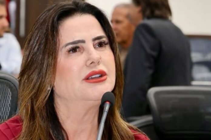 Woman with dark hair and red lipstick speaking into a microphone at a conference table.
