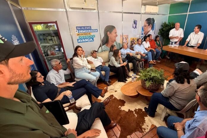 Group of people seated in a semi-circle listening to two men speaking at a long table during a community meeting, posters on the wall.