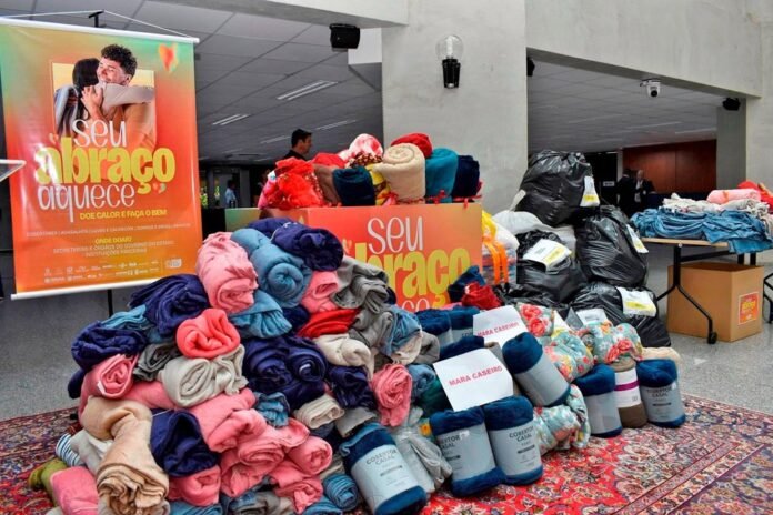 Pile of colorful blankets and throws stacked on a rug next to donation signs that read 'seu abraço' inside a building lobby. Large black trash bags and labeled boxes are visible in the background.