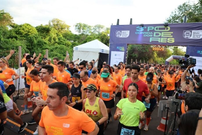Runners in bright orange shirts burst from the starting line under a purple arch reading Corrida do Res, with trees and tents in the background.