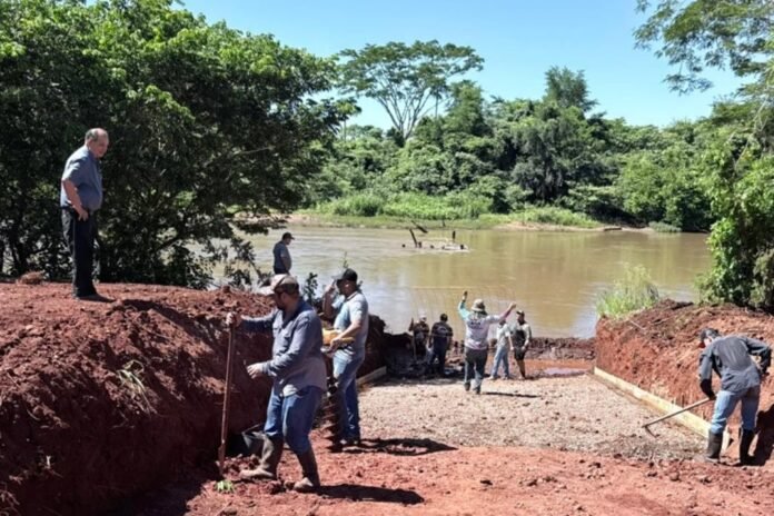 A group of workers dig and move red soil along a riverbank while others stand on the edge, preparing a construction site near the water.